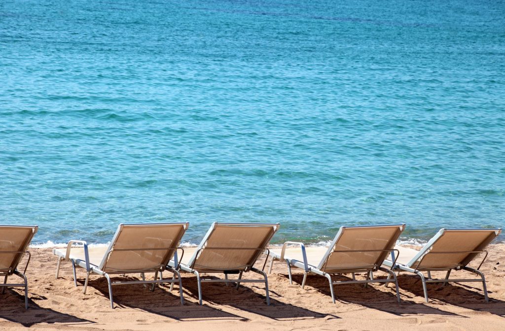 Row of beige beach loungers on a sandy shore facing clear turquoise sea.