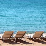 Row of beige beach loungers on a sandy shore facing clear turquoise sea.